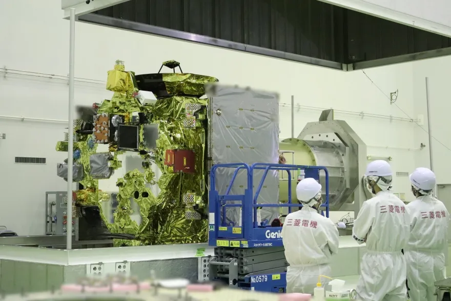Technicians in white cleanroom suits inspect a gold-foil-wrapped spacecraft beside a blue lift in a bright laboratory, conveying careful precision.