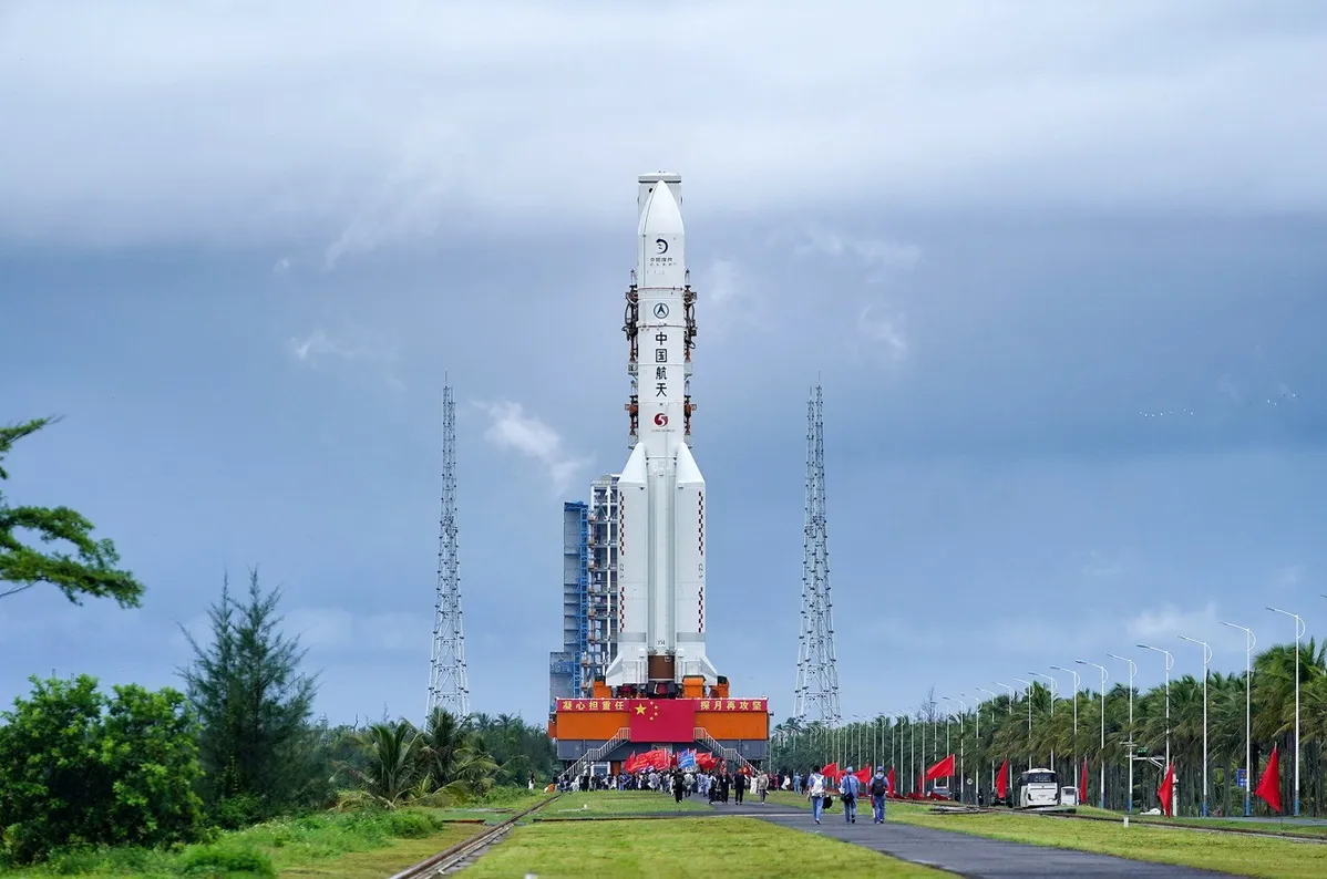 a rocket sitting at launching pad, with bright blue sky in the backgound. there is china flag infront of the rocket hanging down with longshot that show the green area of the place.